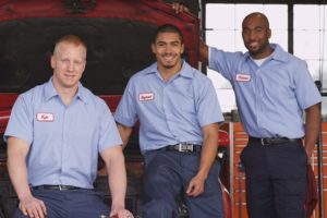 Three auto shop mechanics wearing blue uniforms and standing in front of a car