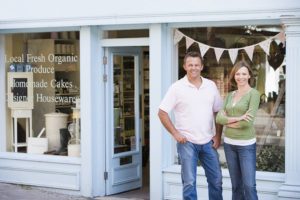 Smiling man and woman standing in front of their local shop that sells produce, cakes and housewares.
